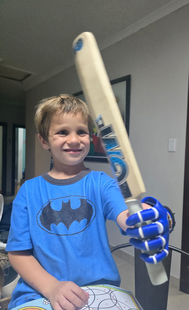Noah grinning and holding a cricket bat up with his blue prosthetic hand, looking proud and happy