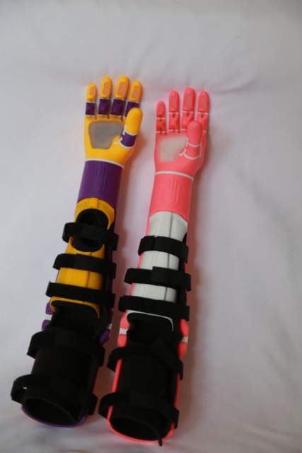 Both completed Kinetic Arms standing upright side by side on white fabric, fingers open, pink on the left and purple/yellow on the right
