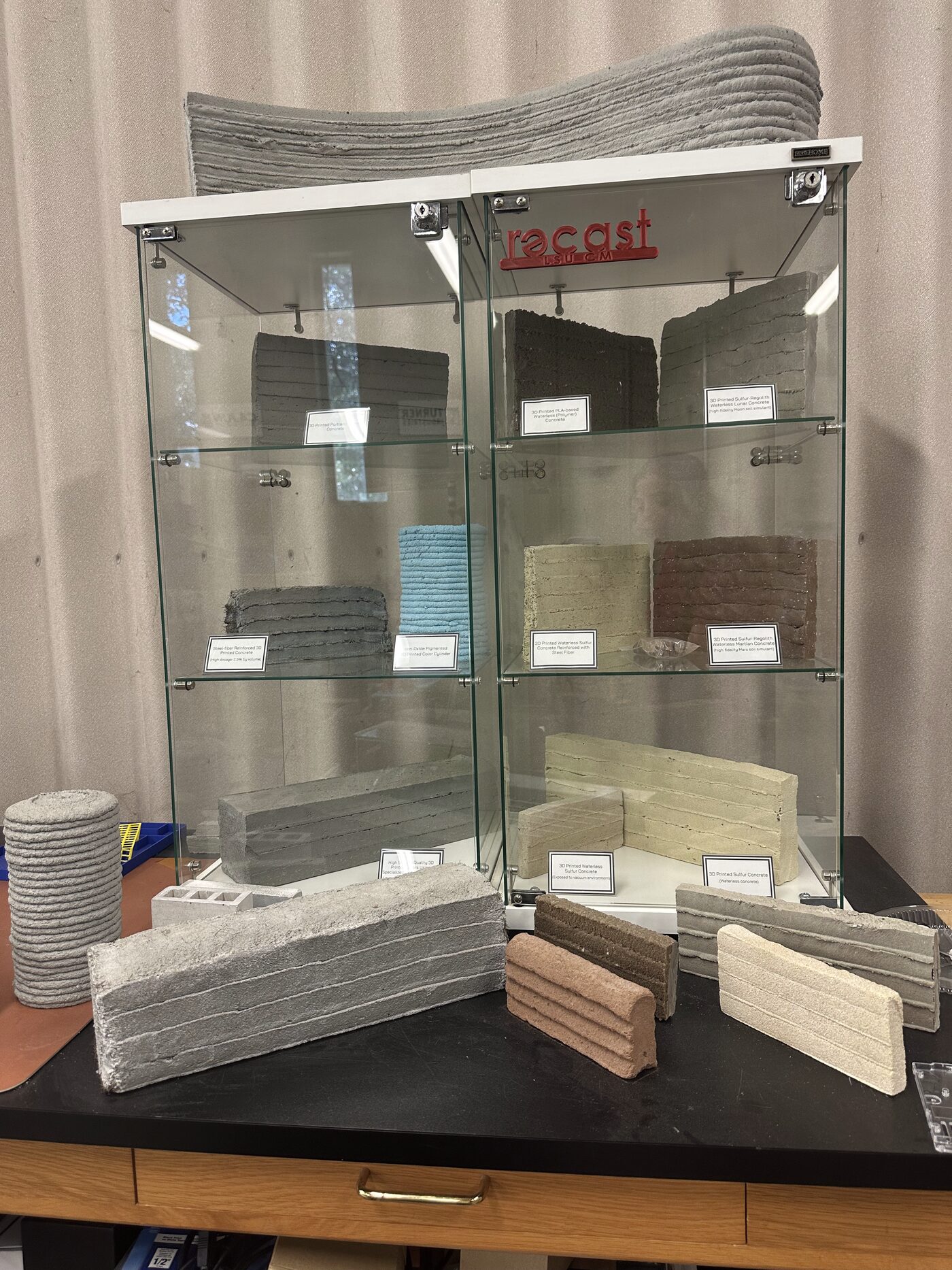 Display cabinet at the RECAST Lab showing 3D-printed concrete and sulfur wall samples in multiple mixes and colors, with the recast LSU logo