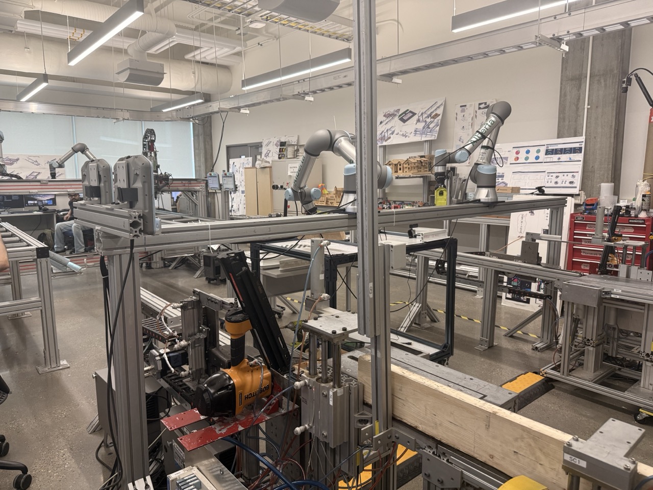 The wood-frame wall assembly machine in the MCRG lab at the University of Alberta, with UR5 robot arms on an overhead gantry above the conveyor stations